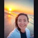 "Smiling woman taking a selfie on a beach at sunrise, wearing a white towel, with wet hair, the sun low over the ocean, and waves gently rolling onto the shore under a colorful sky."
