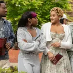 "Four young adults stand together outdoors on a college campus, dressed in casual and semi-formal clothes, holding pamphlets and talking, with trees, other people, and a brick building in the background."