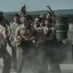 "A group of WWII-era soldiers in dusty uniforms are shouting and cheering around two men, possibly during a brawl or contest, outdoors near metal barrels and mountains in the background."